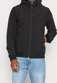 Giacca nera con cappuccio, con chiusura a zip frontale, due tasche laterali e una tasca sul petto con zip. Presenta polsini a costine e un fondo elastico.