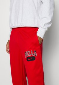 Pantalones de chándal rojos con una cinturilla elástica, bolsillos laterales y un gráfico negro de 'BULLS' encima de un logo de Nike en el muslo izquierdo.