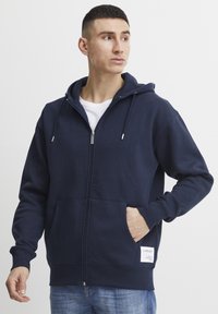 Navy blue zip-up hoodie made of soft material, featuring a spacious front pocket and drawstrings on the hood. White label detail at the hem.