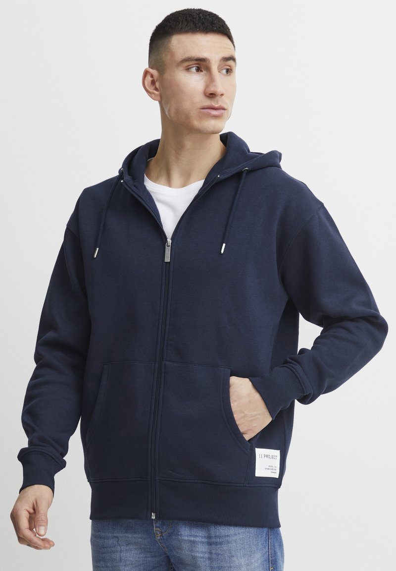 Navy blue zip-up hoodie made of soft material, featuring a spacious front pocket and drawstrings on the hood. White label detail at the hem.