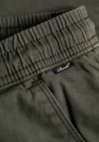 Olivgrüner Stoff mit strukturiertem Oberflächen, ausgestattet mit einem elastischen Bund und einem kleinen schwarzen Etikett mit weißem Text, das in die Naht genäht ist.