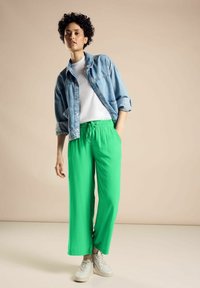 Zielone szerokie spodnie na elastycznej talii w zestawieniu z białą koszulką i dżinsową kurtką. Model nosi białe sneakersy.