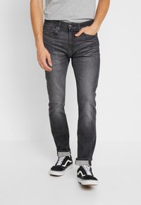 Jeans de mezclilla negra con un corte ajustado, ligero desvanecimiento, cinco bolsillos y una etiqueta con el logo. Combinados con zapatillas bajas en negro y blanco.