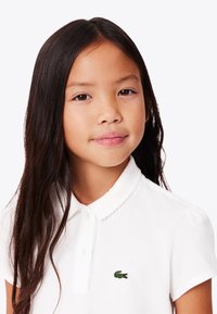 Ung flicka med långt mörkt hår som bär en vit polo t-shirt med en liten grön krokodil-logotyp på bröstet, neutral bakgrund.