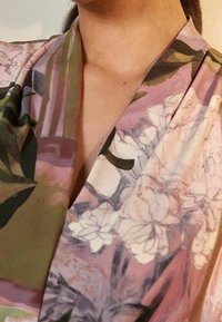 Blomster mønstret stof med bløde teksturer i nuancer af grøn, pink og hvid. Har en dyb V-halsudskæring med draperede detaljer.