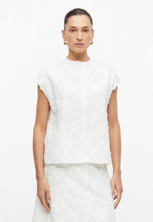 Vrouw met donker haar in een witte mouwloze textuurrijke bloemen top en bijpassende rok, draagt gouden oorringen, staand tegen een witte achtergrond.