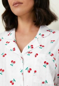 Witte shirt met een kraag, voorzien van rode kersjes borduursel en groene stelen. Inclusief een borstzak en een knoopsluiting aan de voorkant.
