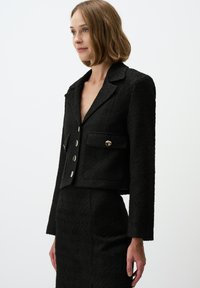 Schwarze, kurze Blazerjacke aus strukturiertem Stoff, mit Reverskragen, zwei vorderen Taschen und silbernen Knöpfen. Kombiniert mit einem passenden Rock aus strukturiertem Material.