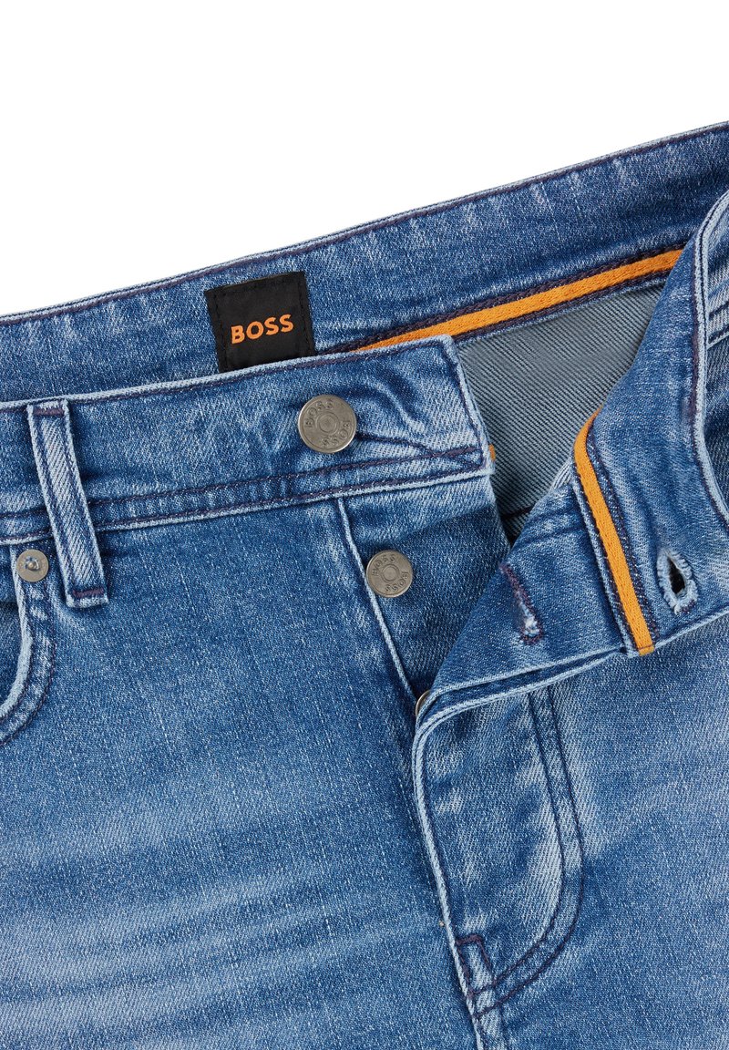 Jean en denim bleu clair avec un aspect usé, comportant une étiquette noire "BOSS", des éléments en métal argenté et un accent orange à la ceinture intérieure.