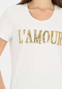 Biała koszulka z krótkim rękawem, ozdobiona złotymi cekinami ułożonymi w napis "L'AMOUR", z zaokrąglonym dekoltem i gładką bawełnianą teksturą.