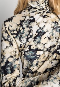 Langarm turtleneck gemaakt van zachte stof, met een meerkleurig abstract patroon in beige, zwart en teal, met verschillende texturen.