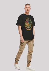 Black t-shirt featuring a detailed multicolored crest design, paired with beige cargo pants and white sneakers. Simple, casual look.