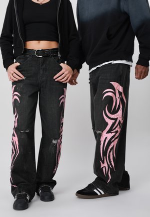 Twee personen dragen zwarte jeans met wijd uitlopende pijpen en roze tribal ontwerpen, zwarte sneakers en donkere bovenkleding, staand tegen een lichte achtergrond.