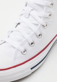Weiße Leinenschuhe mit Metallösen, flachen weißen Schnürsenkeln, Gummispitze und roten sowie blauen gestreiften Sohlenakzenten.