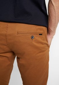 Brown cotton pants with a back pocket featuring a black button, textured fabric, and a branded tag on the side.