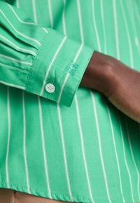 Chemise verte à manches longues avec des rayures verticales blanches, un poignet boutonné et un détail brodé sur le poignet d'une personne à la peau foncée.