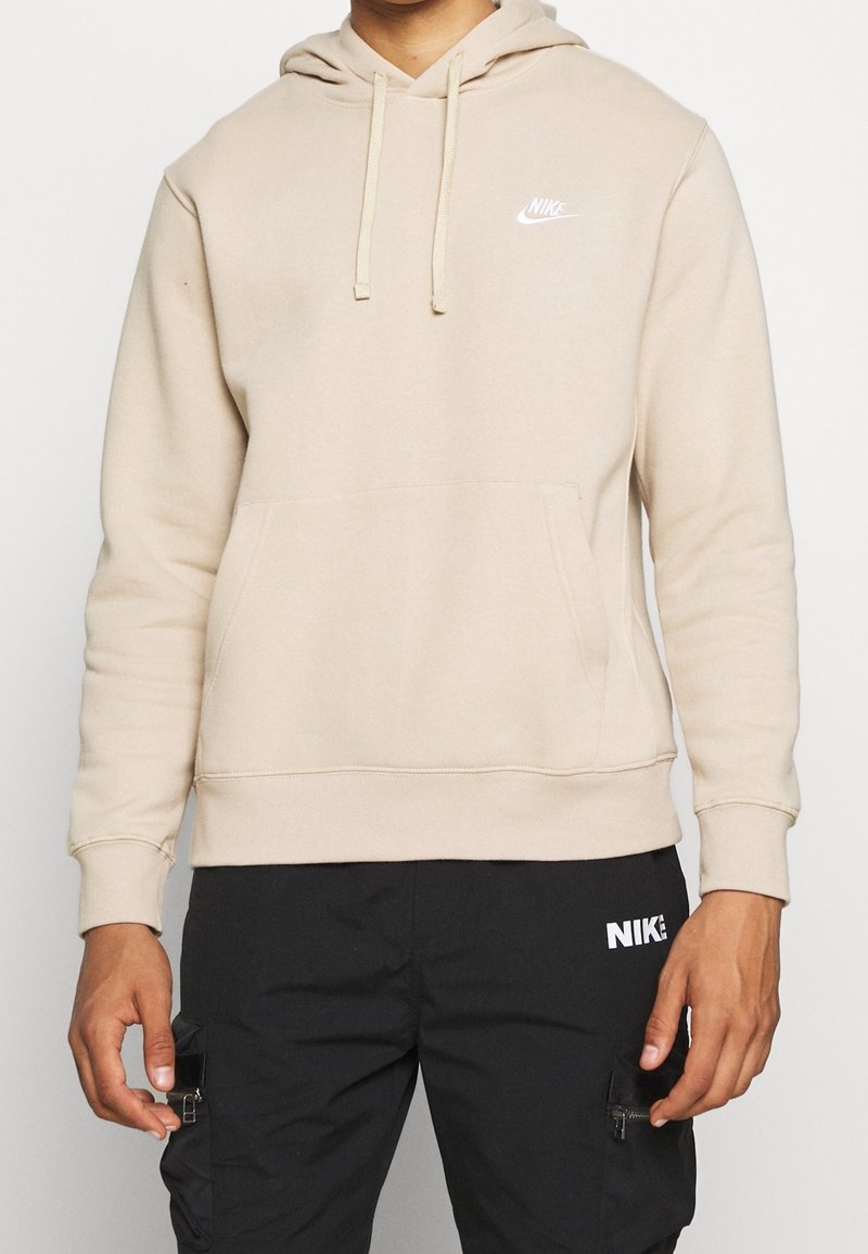 Sweat à capuche beige Nike avec poche avant, porté avec un pantalon noir Nike avec poches cargo zippées.