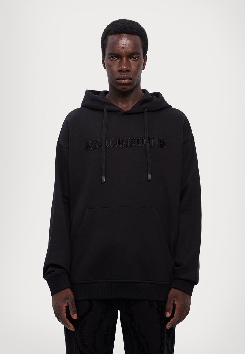 Sudadera negra con un bolsillo canguro en la parte frontal, texto en relieve negro "VERSACE JEANS COUTURE" en el pecho, capucha ajustable con cordón, puños acanalados.