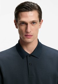 Ciemnogranatowa koszulka polo z teksturowanej tkaniny, z krótkimi rękawami i klasycznym kołnierzykiem. Z przodu znajdują się trzy czarne guziki.
