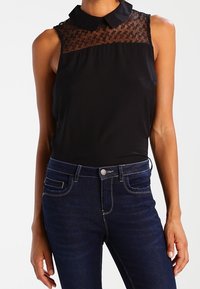 Haut noir sans manches avec un empiècement en dentelle et un col classique, associé à un jean en denim bleu foncé avec une conception à cinq poches et des coutures claires.