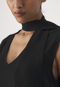 Μαύρο χωρίς μανίκια ένδυμα με V-λαιμό και λεπτομέρεια προσαρμοσμένου λαιμού. Λείο ύφασμα με ματ φινίρισμα, μινιμαλιστικός σχεδιασμός.