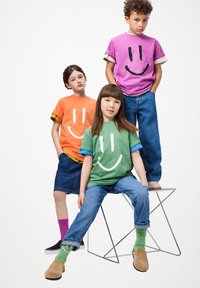 Tres niños que llevan camisetas de caras sonrientes coloridas, jeans y faldas, posando frente a un fondo blanco liso.