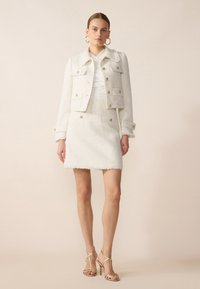 Giacca in tweed bianco con quattro tasche e bottoni dorati, abbinata a una gonna coordinata con orlo frangiato, completata da sandali con tacchi a strappo dorati.