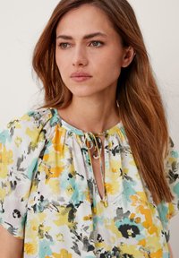 Eine Frau mit langen braunen Haaren, die eine weiße Bluse mit gelben, türkisfarbenen und schwarzen Blumenmustern sowie einem gebundenen Ausschnitt trägt und leicht zur Seite schaut.