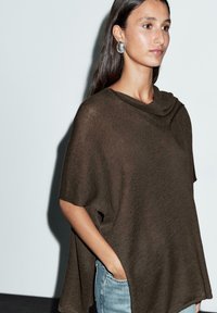 Brun stickad poncho med draperad halsringning, vida ärmar och sidouppskärningar. Tillsammans med ljusblå denimjeans. Slät textur, avslappnad passform.