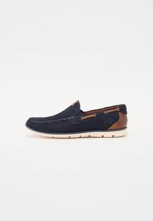 Mørkeblå ruskinds slip-on sko med beige sål; har kontrastsyninger, brun læderdetalje på hælen og dekorative sidehuller.