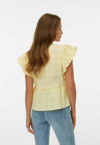 Lichtgele blouse met gerimpelde mouwen, gestructureerde stof en een peplum taille. Gecombineerd met lichtblauwe strakke jeans.