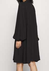 Svart klänning med långa puffärmar, rund halsringning och en fladdrande silhuett. Tyget verkar mjukt med subtila rynkor bak.