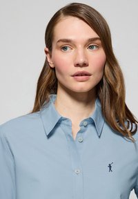 Ljusblå skjorta med krage och tryckknappdesign, med en liten broderad logotyp på vänster bröst och en slät tygstruktur.