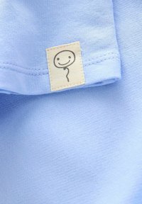 Tessuto azzurro chiaro con una superficie liscia, dotato di un piccolo etichetta color crema con un semplice design di volto sorridente.
