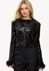 Veste courte noire à sequins avec manches longues, dotée d'une fermeture à boutons sur le devant et d'accents en fausse fourrure noire sur les poignets. Texture lisse et brillante.