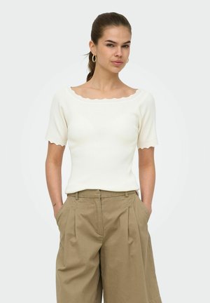 Femme aux cheveux bruns en queue de cheval, portant des boucles d'oreilles en forme d’anneaux dorés, un haut blanc à manches courtes avec des bords festonnés, et un pantalon ample beige avec les mains dans les poches.