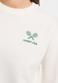 Sweatshirt couleur crème avec un col rond côtelé, présentant un graphique vert de raquettes de tennis croisées et le texte "racket club" en dessous.