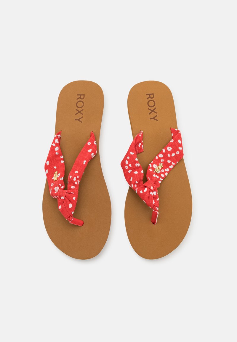 Red fabric flip-flops featuring floral patterns, knotted straps, and brown textured soles with "ROXY" branding embossed on them.