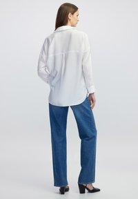 Witte overhemd met lange mouwen, relaxte pasvorm en ronde onderkant; gecombineerd met losse blauwe denim jeans en zwarte blockhakken.