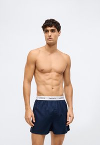 Tânăr cu păr creț și închis la culoare, fără tricou, purtând boxeri Lacoste de culoare albastru închis cu un brâu alb cu sigla, stând în fața unui fundal simplu.