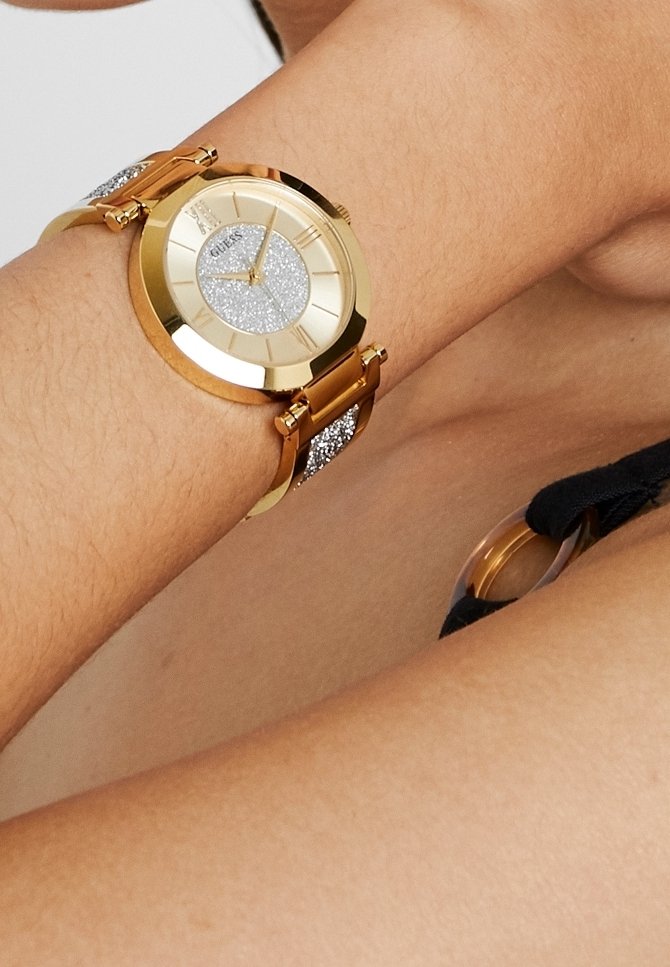 Montre-bracelet en or avec un cadran argenté scintillant et des chiffres romains sur l'avant-bras d'une personne portant un vêtement à sangle noire.