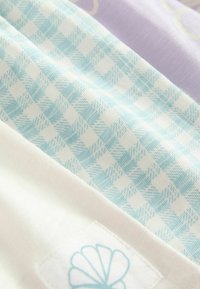 Baumwollstoff im hellblauen Karomuster mit cremefarbenen und sanft lavendelfarbenen Abschnitten. Auffälliges Muschel-Logo auf einem weißen Stoffstück. Glatte Textur.