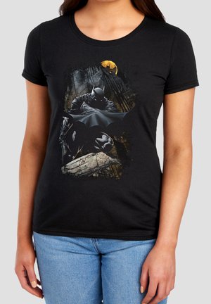 Person trägt ein schwarzes T-Shirt mit Batman-Grafik, die Batman zeigt, wie er auf einem Dach unter einem gelben Mond in einer dunklen Stadtkulisse kauert.