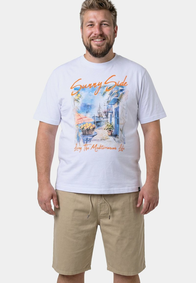 Hombre sonriente vistiendo una camiseta blanca con estampado colorido de una calle mediterránea y un barco, y pantalones cortos color caqui, de pie frente a un fondo liso.