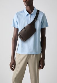 Bolso de cuero marrón con un acabado texturizado, que cuenta con compartimentos con cremallera y una correa ajustable, llevado sobre una camisa polo de color azul claro.