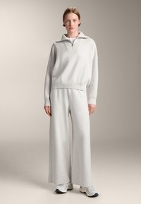Hellgrauer Sweatshirt mit einem halben Reißverschluss-Kragen, kombiniert mit grauen weiten Hosen. Beide bestehen aus weichem, strukturiertem Stoff. Getragen mit weißen Sneakers.