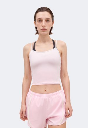 Persoană tânără cu păr scurt și ud poartă un top scurt roz deschis peste un sutien sport negru și pantaloni scurți atletici roz deschis asortați.