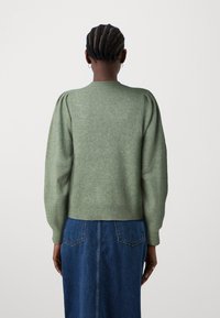 Zelen tretiran pulover s nabranimi rukavi, rebrastim robom in sproščenim krojem, nošen preko jeanskega krila. Mehka tekstura in preprost dizajn.