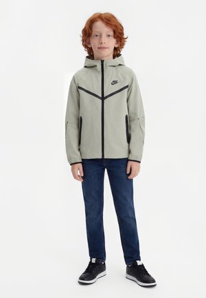 Rude dziecko stoi w jasnoszarej kurtce z kapturem Nike, ciemnoniebieskich dżinsach i czarnych trampkach na jednolitym białym tle.