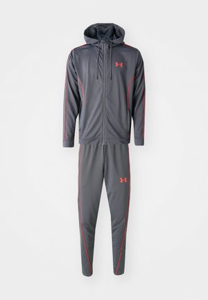 Survêtement de sport pour homme en tissu gris, comprenant une veste à capuche avec des accents rouges et un pantalon fuselé assorti. Logo affiché de manière proéminente.